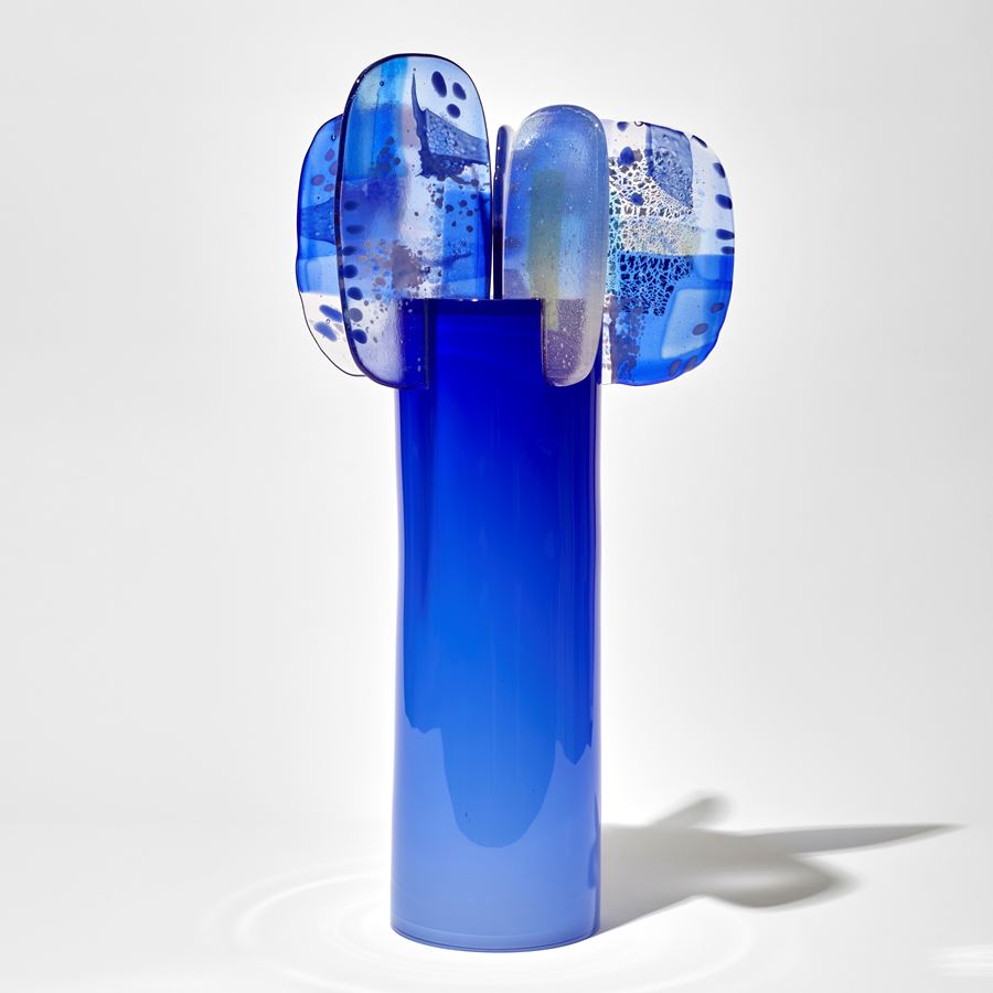 shiny blue cylinder fading from light to darker intensity of colour at the top with five petals perched on the top with abstract patterns and dots in blue and lilac hand made from blown and fused glass 